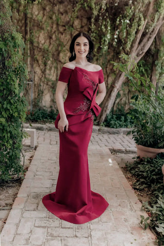 Woman in a wine evening gown with floral embroidery standing outdoors on a stone path.