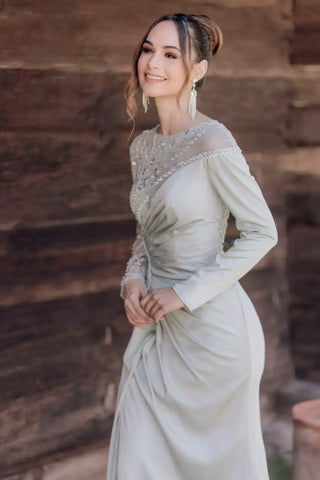 Woman in a light-colored dress with intricate detailing standing against a stone wall.