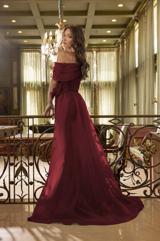 Woman in a burgundy off-shoulder gown standing in an elegant interior setting.