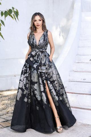 Woman wearing a black and gray floral evening gown with a slit, standing against a white curtain.