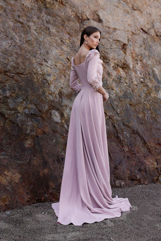 Woman in a long, light purple dress standing against a rocky wall.