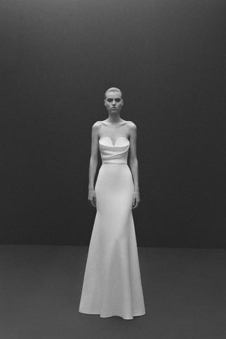 Model wearing the Alex Perry Hartley wedding dress, featuring a strapless sweetheart neckline, elegant drape detail across the bust, and a form-fitting silhouette in satin crepe fabric. The gown is complemented by an extended train and a contrast satin belt, showcased against a minimalist dark background, highlighting the luxurious and modern design.