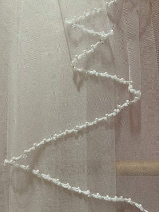 A white bridal veil with a beaded edge displayed on a wooden hanger.