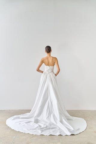 Woman wearing a white strapless wedding dress with a long train on a plain background