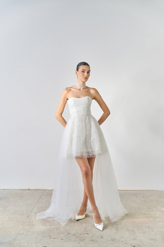 Woman wearing a strapless white lace wedding dress with a long train on a plain background